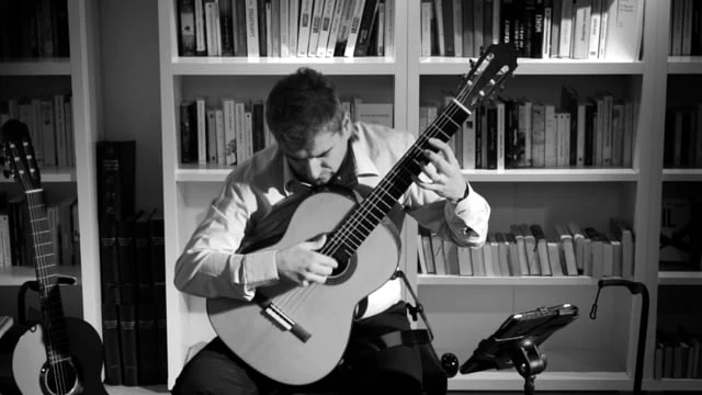 Alexandre Bernoud playing guitar in front of bookshelves, in black and white